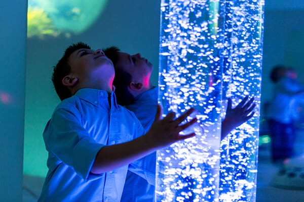 Un enfant regarde les bulles monter dans un tube vitré qui prend place au milieu de la zone de jeux sensorielle, pour jouer dans le calme ou pour se ressourcer quelques minutes.